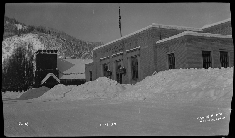 Photo of the Wallace Post Office building in Wallace, Idaho. There are several large piles of snow in front of the building from the road being plowed, and it appears that the snow is blocking the entrance to the building. There is a flag pole with an American flag on the top of the building.