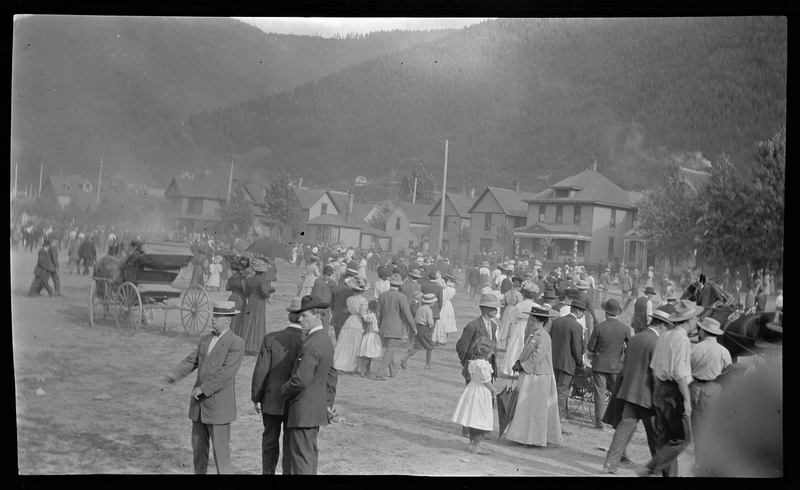 Several large groups of unidentified people gather in an open area of Wallace, Idaho for Roosevelt's visit. Everyone visible are dressed nicely. There are two horse drawn carriages visible, and a line of houses in the background.