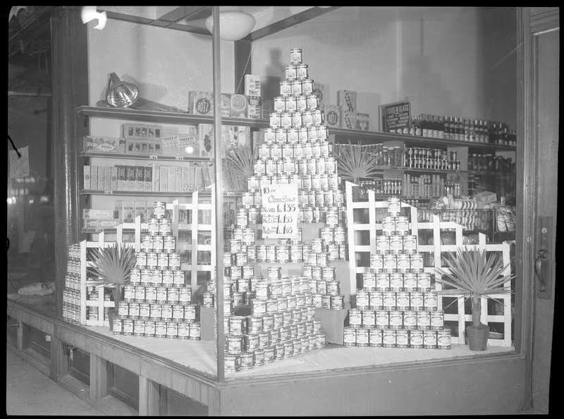 Photo of the window display in Bennet's Store on 5th Street in Wallace, Idaho. The display shows several pyramids made from stacking cants of Roundup Corn, and a sign advertising a sale on the corn.