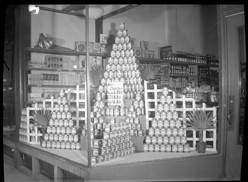 Photo of the window display in Bennet's Store on 5th Street in Wallace, Idaho. The display shows several pyramids made from stacking cants of Roundup Corn, and a sign advertising a sale on the corn.