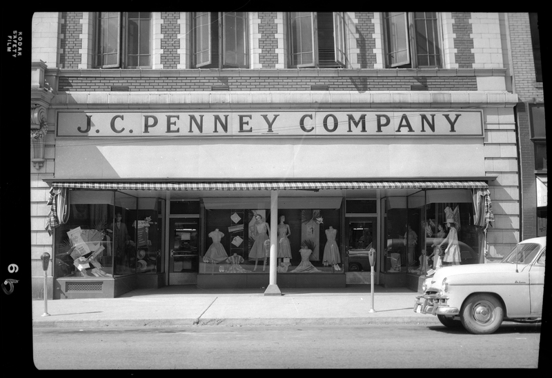 Photo of the storefront of the J. C. Penney Company building. There are several mannequins displaying various clothing items in the front windows, and a car parked on the side of the street next to the building.