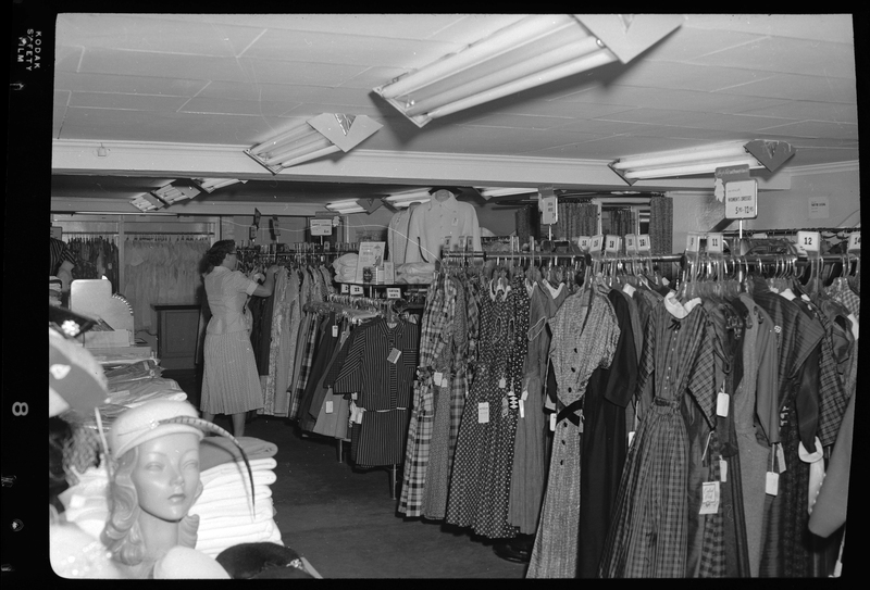 Interior photo of the J. C. Penney Company store. There are several racks of dresses on display, and a woman is actively looking through one of the racks.