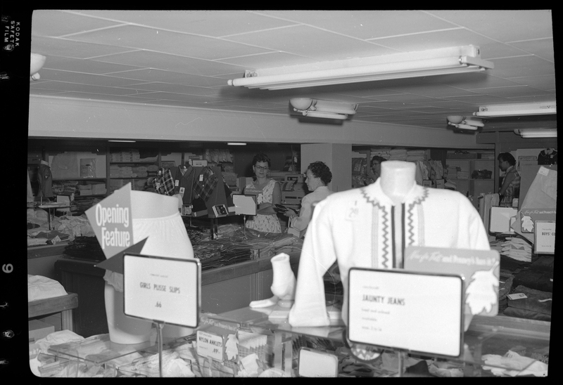 An unidentified woman is checking out at the cash register inside the J. C. Penney Company store. She is partially obscured by clothing displays.