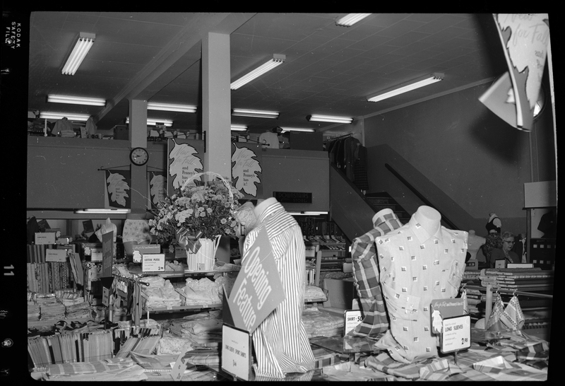 Interior photo of the J. C. Penney Company store. There are several displays of various clothing pictured, including shirts, pants, and lingerie. A woman can be seen shopping in the background.