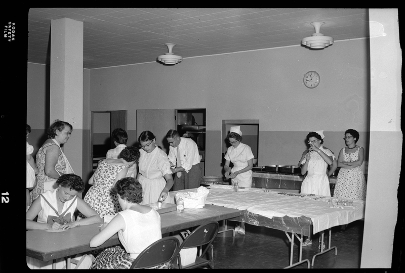 Photo of several unidentified people, mostly women and children, in line at the Shoshone County Polio Shot Clinic getting their vaccines. There are several nurses in the room administering shots. A nurse can be seen prepping a vaccine in a syringe.