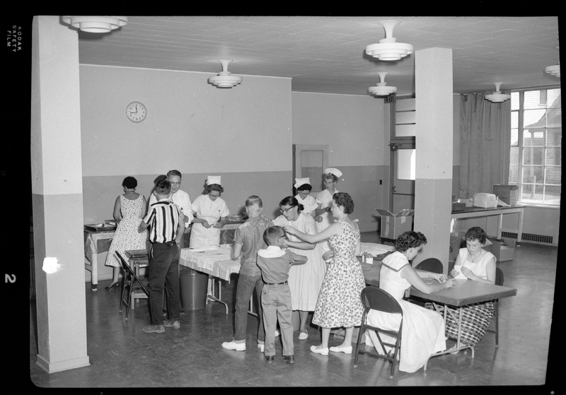 Photo of several unidentified people, mostly women and children, in line at the Shoshone County Polio Shot Clinic getting their vaccines. There are several nurses in the room administering shots.