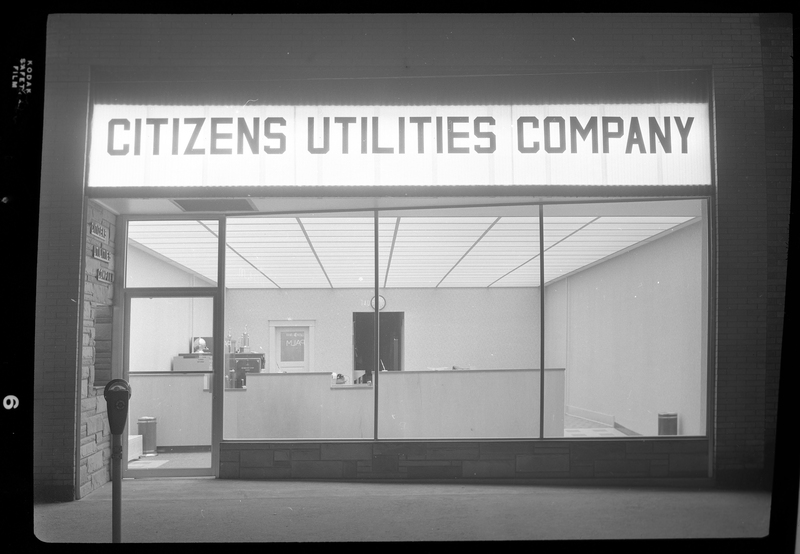 The storefront of Citizens Utility Company. The light of the sign above the entrance and the interior lights are on.