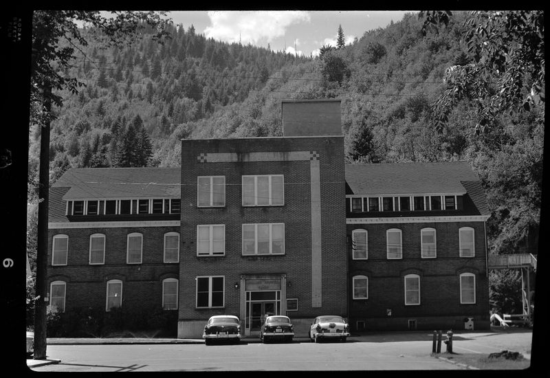 Photo of the front exterior of the Wallace Hospital Building in Wallace, Idaho. The building is at least three stories, and there are three cars parked in front. There is a sign above the main doors that reads, "Wallace Hospital Clinic."