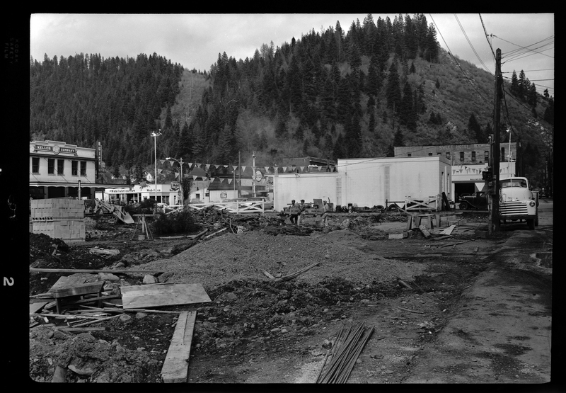 Photo of the ongoing construction process of the Stardust Motel in Wallace, Idaho. The construction side is full of piles of dirt, construction material and equipment, and debris. Two workers are visible on site, and several other businesses are visible in the background.