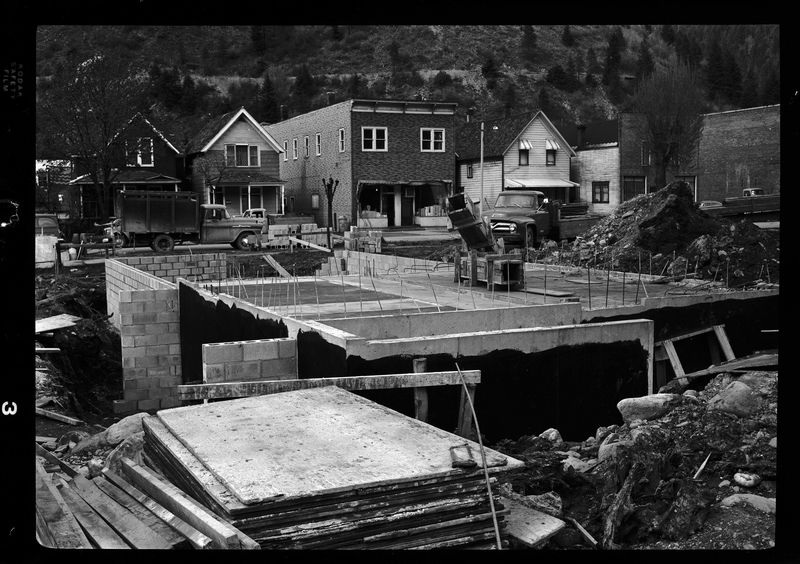 Photo of the foundation for the ongoing construction of a motel in what is likely Wallace, Idaho. The foundation of the building is set within the ground, and some construction above it has been started. There are homes visible in the background.
