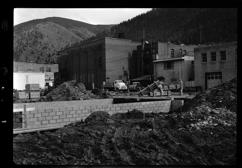 Photo of the foundation for the ongoing construction of a motel in what is likely Wallace, Idaho. The foundation of the building is set within the ground, and some construction above it has been started. Two construction workers are visible on the site that are working near the foundation of the building.