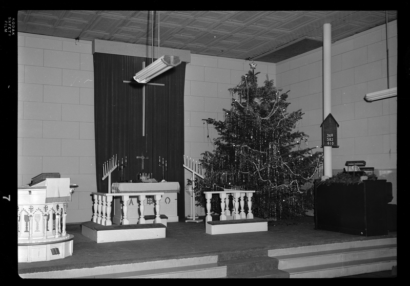 Photo of an altar of an unspecified Lutheran Church in Mullan, Idaho. The altar has a large Christmas tree in one corner that is decorated. The altar itself stands in the middle of the area with candle sticks around it.