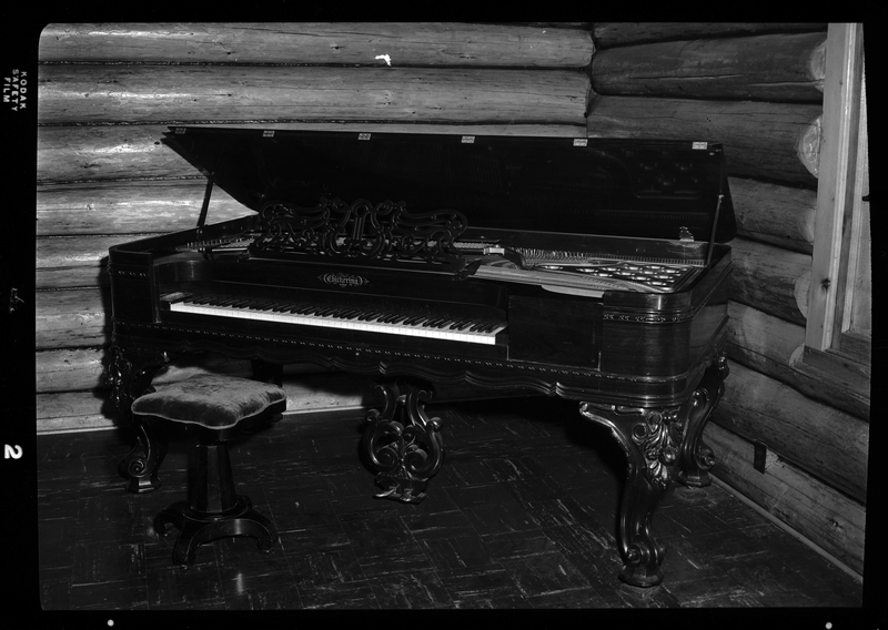 Photo of a Chickering and Sons square grand piano in the Stansfield house. The top of the piano is lifted open and the keys are visible, and there is a single seater piano bench in front of it.