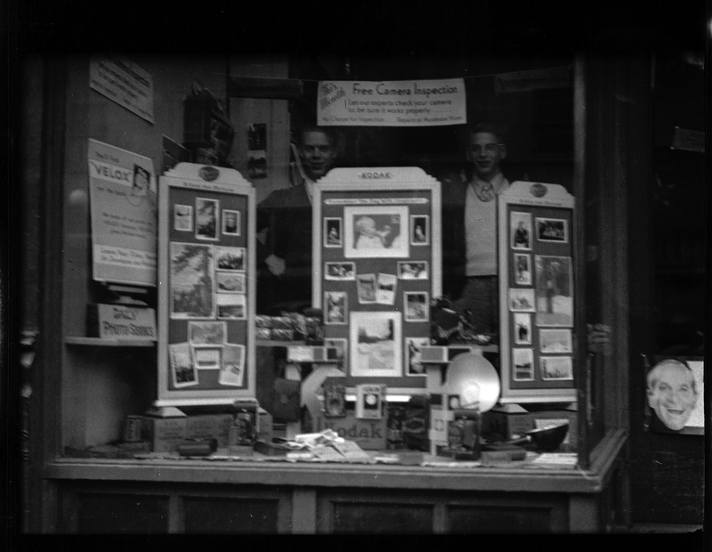 Photo of the camera counter at Tabor's store. There are several displays of photos, cameras, and other photography equipment, and two men stand behind the counter.