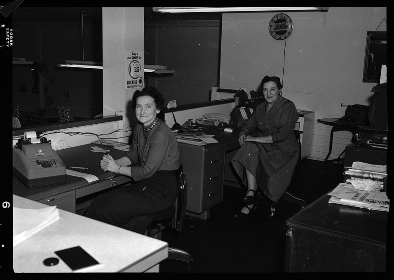Photo of two unidentified women sitting behind individual desks. Both women are turned towards the camera and posing for a photo. Described as "Del Monte Tuna display inside grocery store."