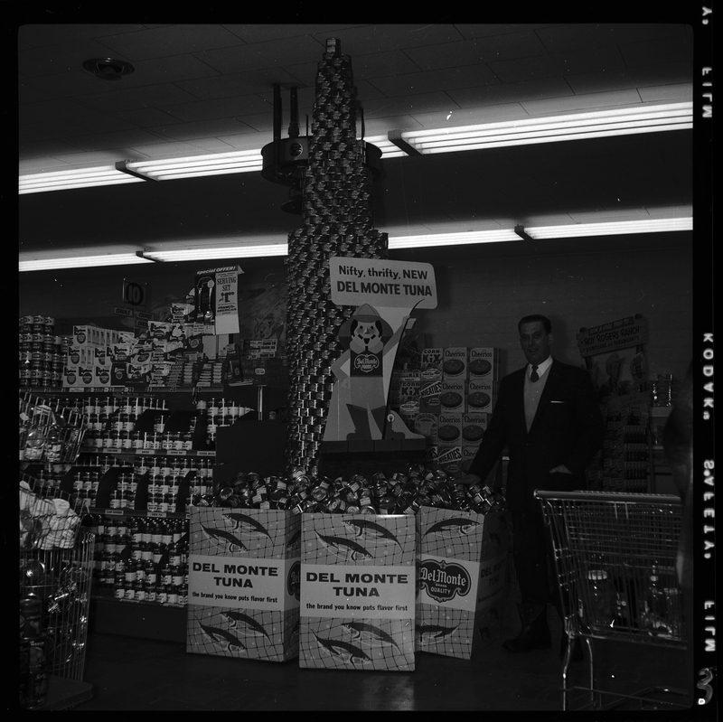 Photo of a man standing next to the Del Monte Tuna display inside of a grocery store. The tuna is stacked in the shape of a tower that nearly reaches the ceiling, and there are tall boxes of the canned tuna surrounding it, and there is a cardboard cutout advertisement for the tuna next to the tower. Other canned and boxed food items are visible in the background.