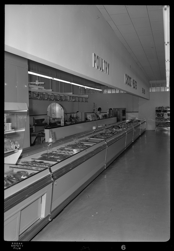 Photo of the poultry counter at the food center. Signs designating the area as the poultry counter are hung on the wall above it, and a row of containers holds food products. Two people are visible working back in the poultry center.