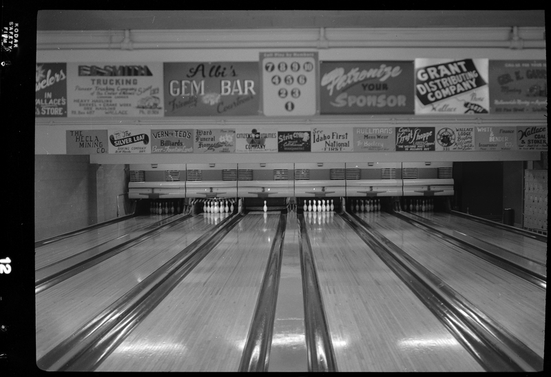Photo of the six bowling lanes at Albi's Bowling Alley. Five of the lanes have all their pins standing, and one lane has just one pin standing. There are poster advertisements all over the walls.