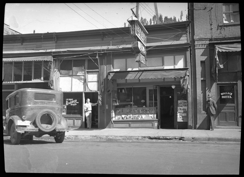 Photo of Les's Barber Shop (and possibly Herman's Jewelry store) next to the Wallace hotel. There is a man standing just outside the door to the barber shop, and another man near the entrance to the hotel. There is a car parked on the side of the road in front of the barber shop.