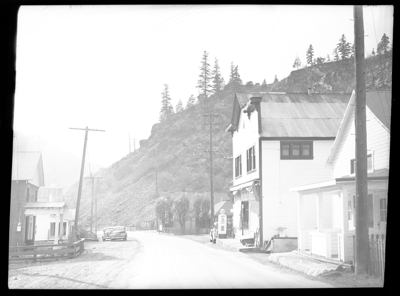 Photo of a street scene in Gem, Idaho. The negative is overexposed and some details are hard to make out, but several buildings, houses, and cars are visible. There is a gas pump besides one of the buildings, and trees are visible on top of the hill next to the buildings.