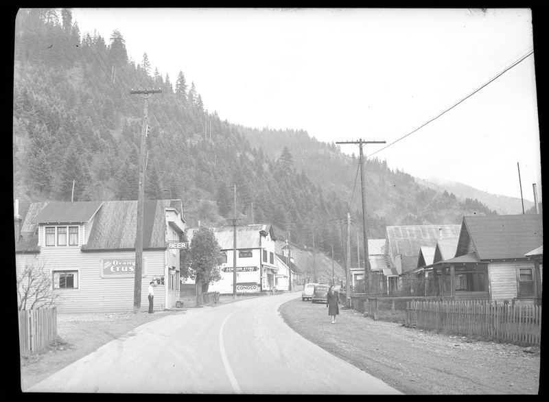 Photo of a street scene in Gem, Idaho. Several buildings and houses line both sides of the street, and a woman can be seen walking towards the photographer. There are two cars parked on the side of the road further down the street, and a man standing next to one of the buildings.