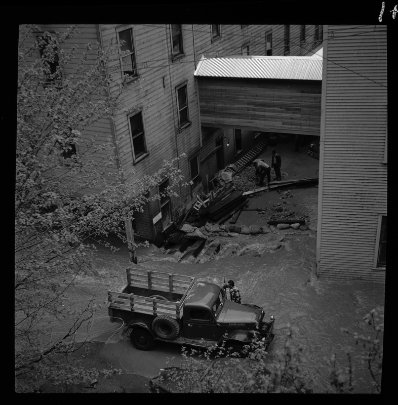 Photo of what is likely a flood in the city of Wallace. High levels of water run near and against buildings, and there are sandbags laid out between the two buildings to stop the water from flooding that way. Two unidentified people stand behind the dryer sandbag area, and another man stands next to a truck which is parked in the rushing water.