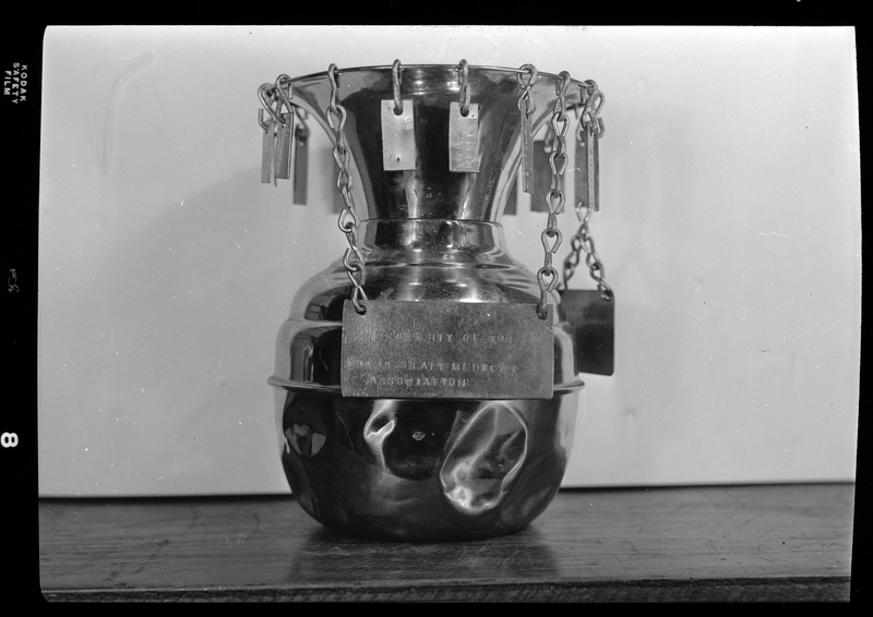 Photo of a metal vase-like item with a sign that reads, "Property of the Idaho State Medical Association." There are hooks around the top of the vase that have metal strips hanging from them. Previously described as "Dr. Paul Ellis Rotating Gaboon."