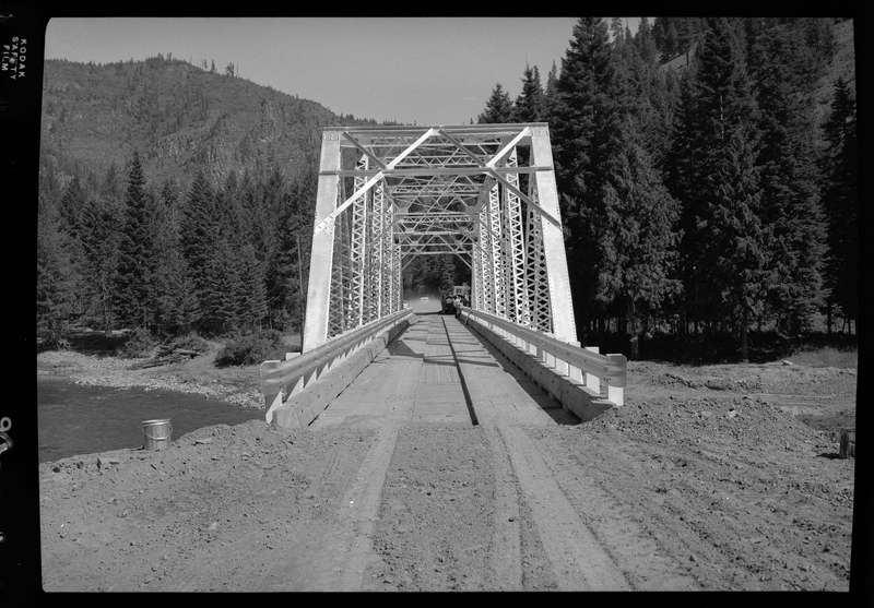 View looking across the Pritchard Bridge in Pritchard, Idaho. Cars and people can be seen on the other side of the bridge, which connects two sides of a river. There are trees surrounding the river and the bridge.