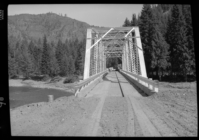 View looking across the Pritchard Bridge in Pritchard, Idaho. Cars and people can be seen on the other side of the bridge, which connects two sides of a river. There are trees surrounding the river and the bridge.