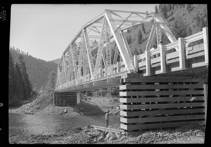 Photo of the ongoing construction of the Pritchard Bridge in Pritchard, Idaho. The bridge frame is in place over the river and settled on the bases. There is a man (presumably a construction worker) standing on the ground under the bridge.