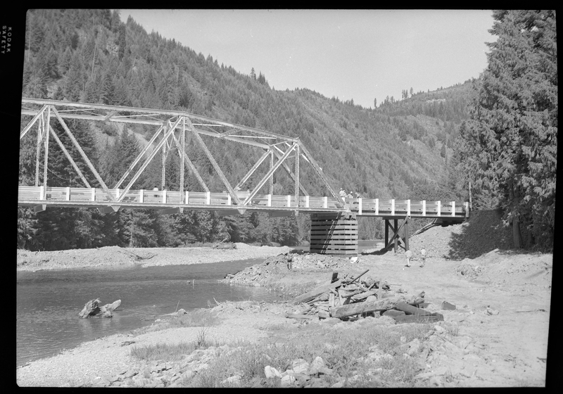 Photo of the ongoing construction of the Pritchard Bridge in Pritchard, Idaho. The bridge frame is in place over the river and settled on the bases. There is a man (presumably a construction worker) standing on the ground under the bridge and more people on the bridge itself.