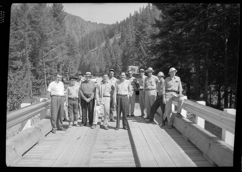 Photo of the construction workers standing on the newly constructed Pritchard Bridge in Pritchard, Idaho. The twelve men stand on the bridge but near one end of it, showing cars and trees behind them.