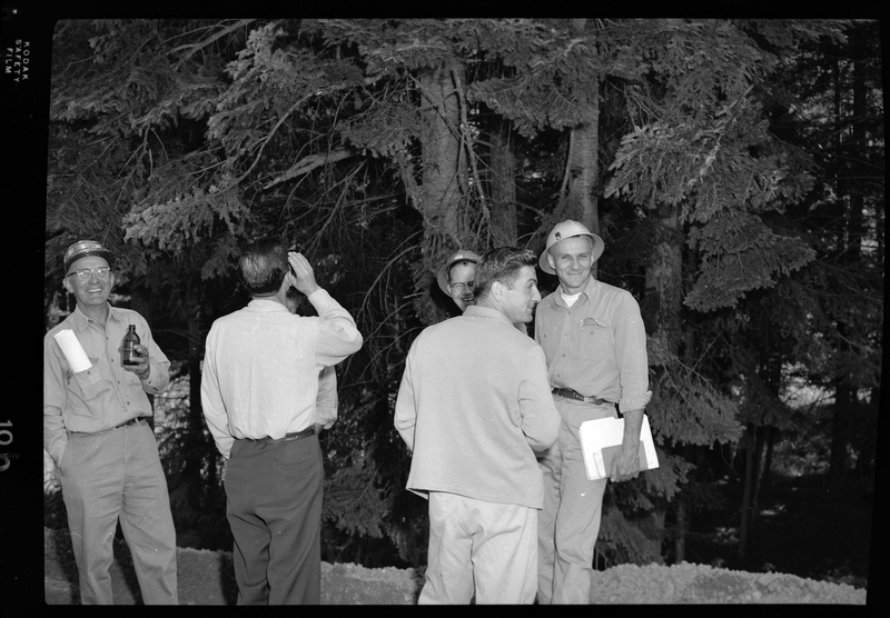 Photo of six of the construction workers from the Pritchard Bridge site talking together. Two of the men have their backs to the photographer, and two of the other men are looking at and smiling at the camera. There are trees directly behind the men.