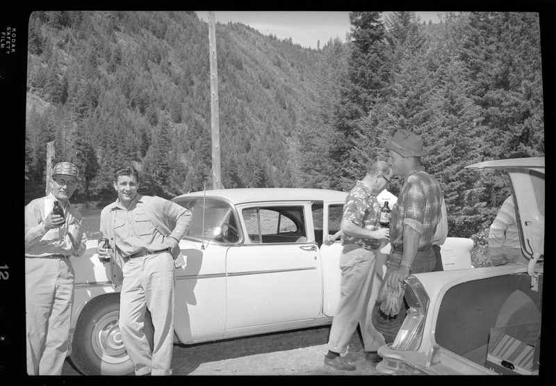 Photo of a few of the construction workers from Pritchard Bridge standing together near their cars and drinking. Two of the men lean against one of the cars and smile at the camera while the other men present are focused elsewhere.