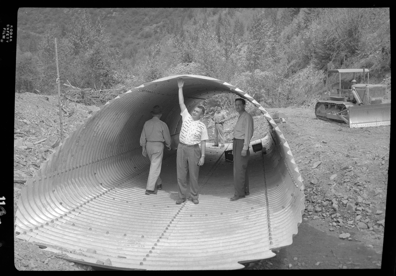 Photo of four construction workers from the Pritchard Bridge site. They are standing inside of an open cylinder of building material, with two of the men posing for the camera (one of which is reaching up to touch the top of the cylinder). One of the other men is faced away and walking back towards the last man, who stands at the other open end of the cylinder.