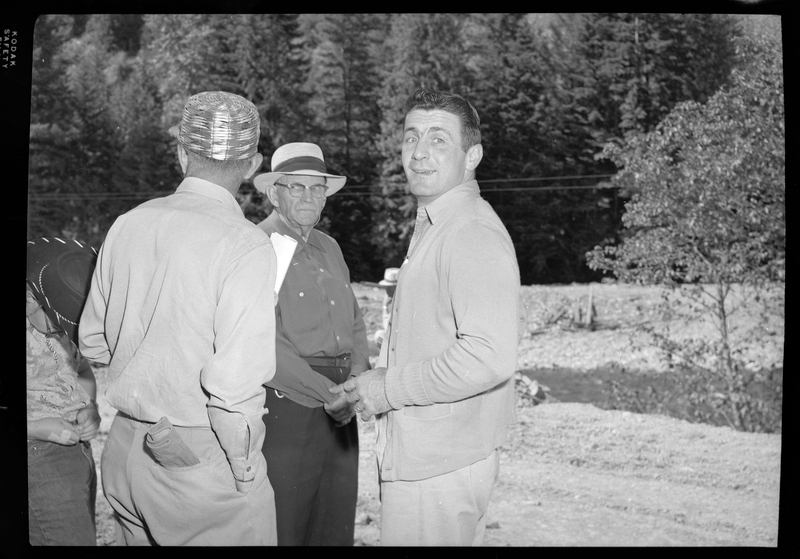 Three of the construction workers from Pritchard Bridge stand together. One of the men is looking over his shoulder and smiling, one man is facing the other but looking the opposite direction, and the third man has his back to the camera. There are trees in the background.