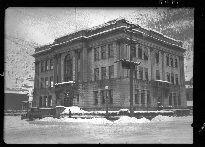 Photo of a Courthouse building in the snow. The snow covered the ground, street, and roof of the building. There is a car parked in front of the building and person walking by it. The building is on the street corner and the photo was taken from the opposite street corner, showing two sides of the building.