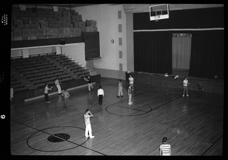 A group of people, likely all men, are playing a game of basketball at the Civic Auditorium in Wallace, Idaho. They are all wearing street clothes, not uniforms. A man is actively jumping up to dunk the ball in the hoop.