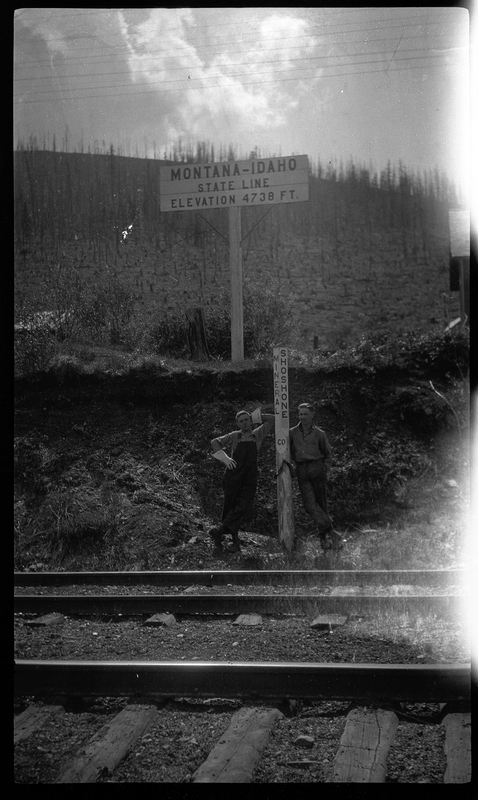Photo of the Montana-Idaho state line road sign. Two men stand underneath it next to another sign that has "Shoshone" on one side and "Mineral" on the other which is likely the sign designating the counties. The state line sign reads, "Montana-Idaho; State Line; Elevation 4738 ft."