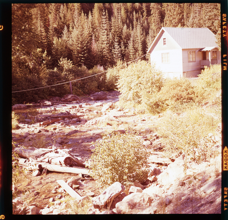 Color photo of a creek that runs next to a building that is likely on A. A. M. Arnold's property on Highway 10. The creek has little water in it, and it is surrounded by trees and bushes. There is a house in the background.
