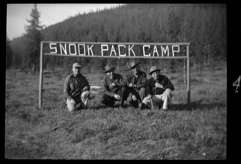 Four men crouch down to kneel underneath the Snook Pack Camp sign. They're all looking at and smiling for the camera.