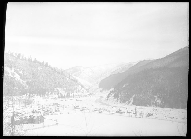 Photo of the city of Silverton, Idaho in the snow. The photo was taken from a higher elevation than the town, capturing most of it. The negative is overexposed and makes the details harder to make out, but several buildings can be made out, as well as the tree covered mountains in the background. Everything is covered in snow.