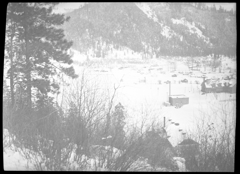Photo of the city of Silverton, Idaho in the snow. The negative is overexposed and not much detail can be made out, but some buildings are visible, as well as the tree covered mountains behind the town. Everything visible is covered in snow.