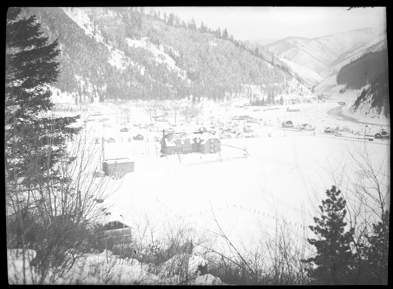 Photo of the city of Silverton, Idaho in the snow. Several houses and buildings are easily visible, all covered in and surrounded by snow. The mountains behind the town are visible and are covered in trees and snow.