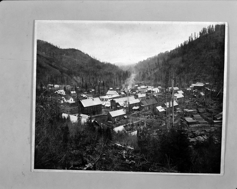 Photo of a photo of the city of Wallace, Idaho taken from a hilltop. There are several buildings centered in the town, and the area to the right has several piles of wooden logs lined up.