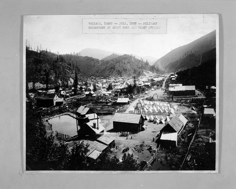 Photo of a photo of a federal encampment in Wallace, Idaho. There is a caption on the photo that reads, "Wallace, Idaho - - July, 1892 - - Military Encampment at about Bank and First streets." The photo shows the military encampment in Wallace Park, which is surrounded by the buildings in city of Wallace.