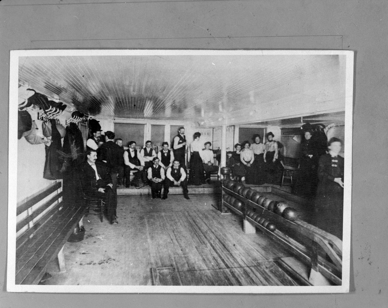 Photo of a photo of several men and women in the bowling alley that was in the basement of Wallace Hotel. The lanes are not visible, but the room appears to only be big enough for two lanes. At least ten people are in the room, and a woman stands in one of the lanes with a bowling ball.