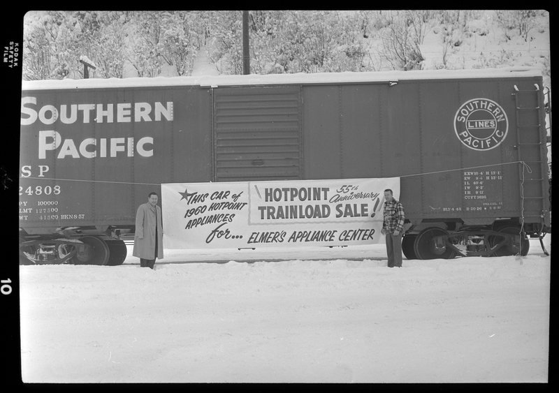 Photo of two men standing on either side of a banner advertisement for Elmer's Appliance Center that is hung across a Southern Pacific train car. The banner reads, "This car of 1960 Hotpoint Appliances for Elmer's Appliance Center; Hotpoint 55th Anniversary Trainload Sale!" There is a heavy layer of snow covering the ground and the top of the train car.