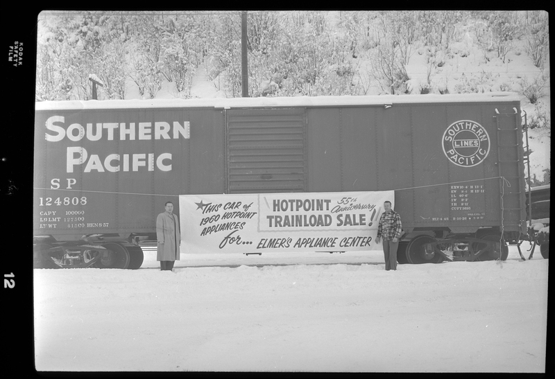 Photo of two men standing on either side of a banner advertisement for Elmer's Appliance Center that is hung across a Southern Pacific train car. The banner reads, "This car of 1960 Hotpoint Appliances for Elmer's Appliance Center; Hotpoint 55th Anniversary Trainload Sale!" There is a heavy layer of snow covering the ground and the top of the train car.