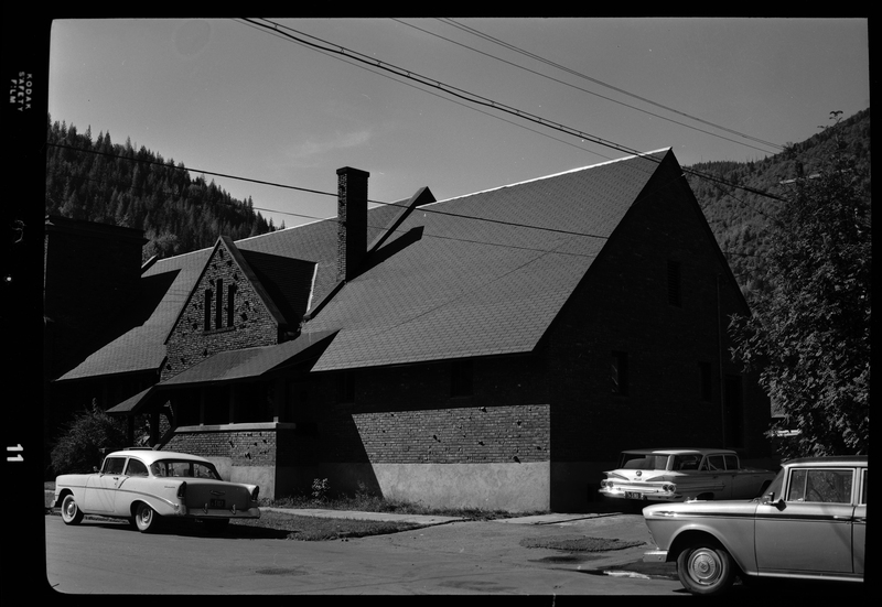 Photo of the exterior of an Episcopal Church in Wallace, Idaho. There are several cars parked along the road and in the alley next to the church.